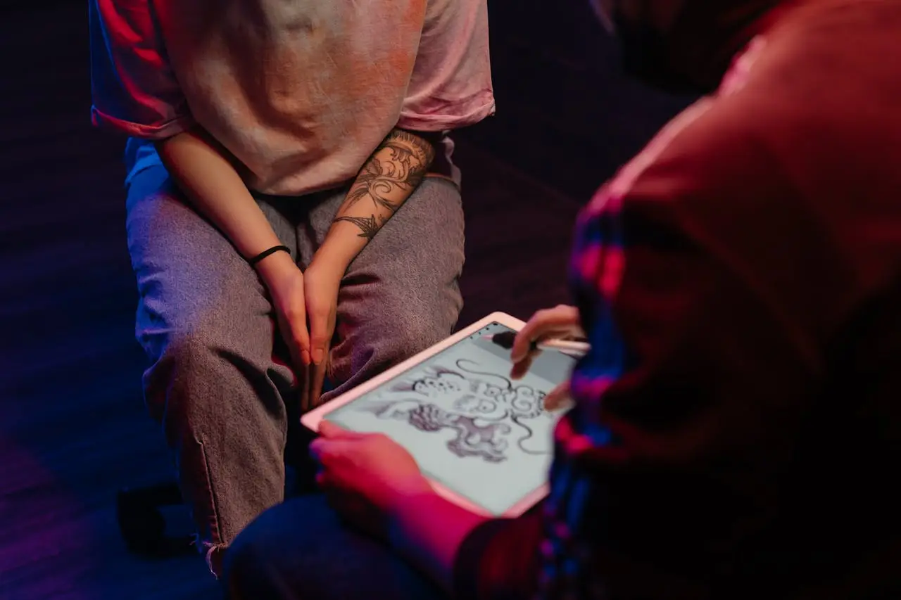 A tattoo artist and client discussing a dragon design on a digital tablet in a dimly lit tattoo studio.